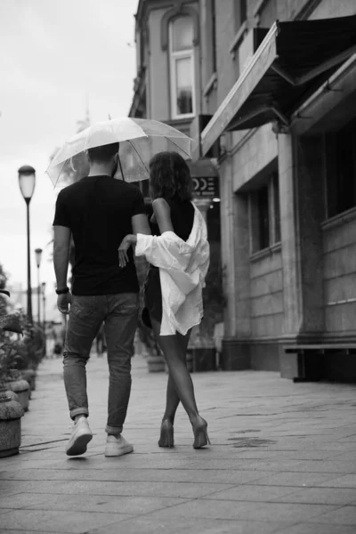 Young couple with umbrella enjoying time together under rain on city street, back view. Black and white effect