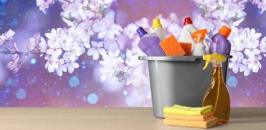 Spring cleaning. Bucket with detergents and tools on wooden surface against blossoming tree, space for text. Banner design clipart