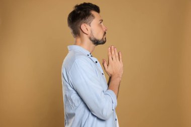 Ellerini kenetleyip bej arka planda dua eden bir adam.