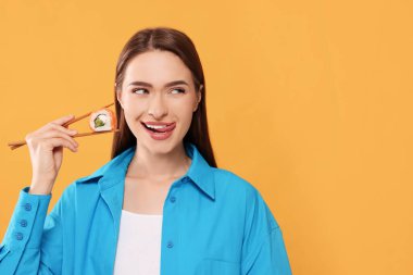 Turuncu arka planda yemek çubuklarıyla suşi dürümü tutan mutlu genç bir kadın. Metin için boşluk