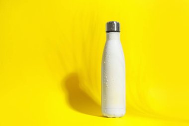 Stylish white thermo bottle with water drops on yellow background, space for text