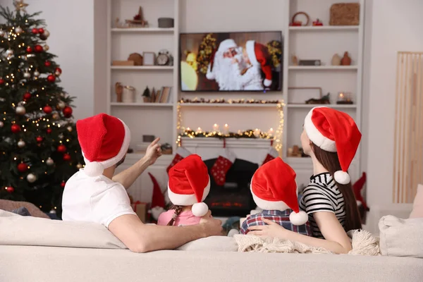 Family wearing Christmas hats watching TV in cosy room, back view. Winter holidays atmosphere