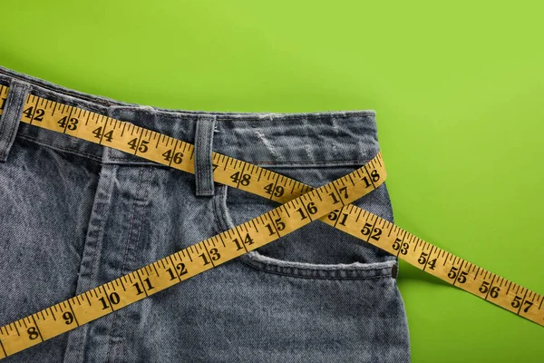 Jeans with measuring tape on green background, top view. Weight loss concept
