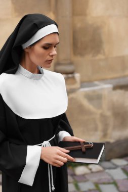 Young nun with Christian cross and Bible near building outdoors