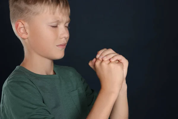 Boy with clasped hands praying on black background, space for text