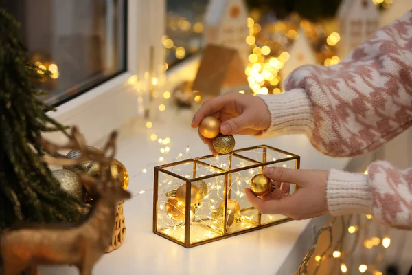 Christmas atmosphere. Woman putting beautiful baubles into decorative container on window sill indoors, closeup