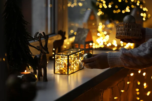 Christmas atmosphere. Woman holding beautiful baubles near window sill indoors, closeup
