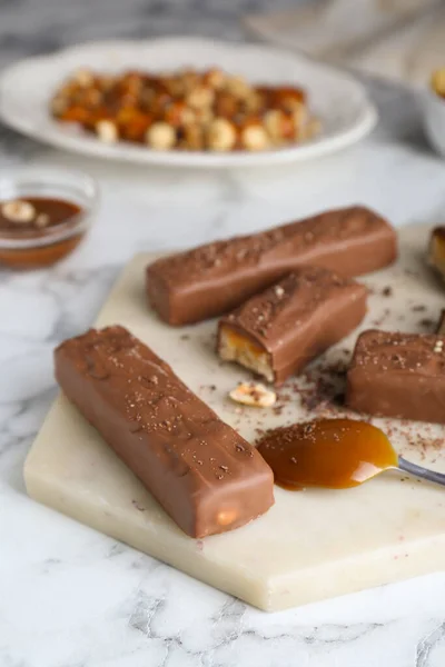 Board with delicious chocolate bars, caramel and nuts on white marble table, closeup