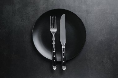 Ceramic plate, fork and knife on black table, top view
