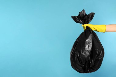 Janitor in rubber glove holding trash bag full of garbage on light blue background, closeup. Space for text
