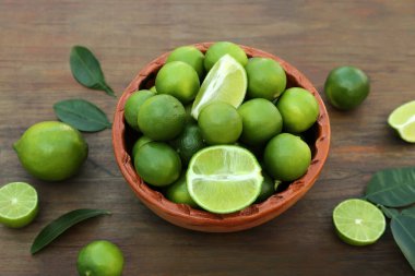 Whole and cut fresh ripe limes with green leaves in bowl on wooden table