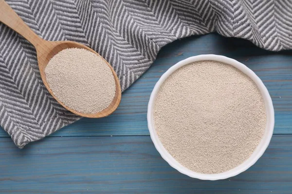 Bowl and spoon with active dry yeast on light blue wooden table, top view