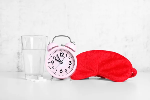 Red sleep mask, glass of water, pills and alarm clock on white table