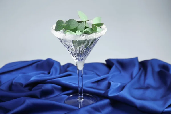 Beautiful martini glass with sugar rim and eucalyptus leaves on blue satin against light grey background