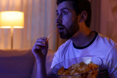 Man eating chips while watching TV on sofa at night. Bad habit