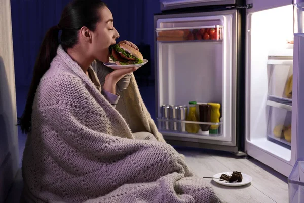 Young woman eating burger near refrigerator in kitchen at night. Bad habit
