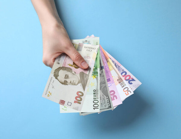 Currency exchange. Woman holding euro, hryvnia and dollar banknotes on light blue background, closeup