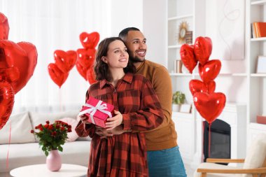 Happy couple celebrating Valentine's day. Beloved woman with gift box in room decorated with heart shaped air balloons