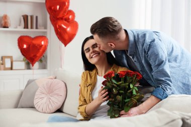 Boyfriend presenting beautiful bouquet of roses to his girlfriend in room decorated with heart shaped balloons. Valentine's day celebration