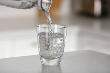 Pouring water from bottle into glass on light grey marble table in kitchen. Space for text