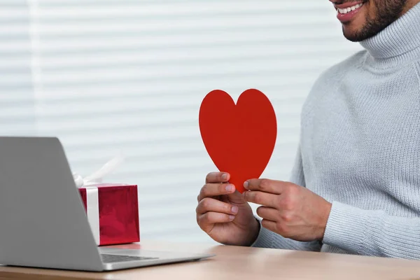 Valentine\'s day celebration in long distance relationship. Man holding red paper heart while having video chat with his girlfriend via laptop, closeup