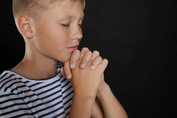 Boy with clasped hands praying on black background, closeup. Space for text