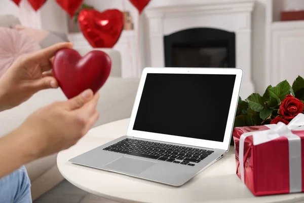 Valentine's day celebration in long distance relationship. Woman holding red wooden heart while having video chat with her boyfriend via laptop indoors, closeup