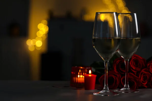 Glasses of white wine, burning candles and rose flowers on grey table against blurred lights, space for text. Romantic atmosphere