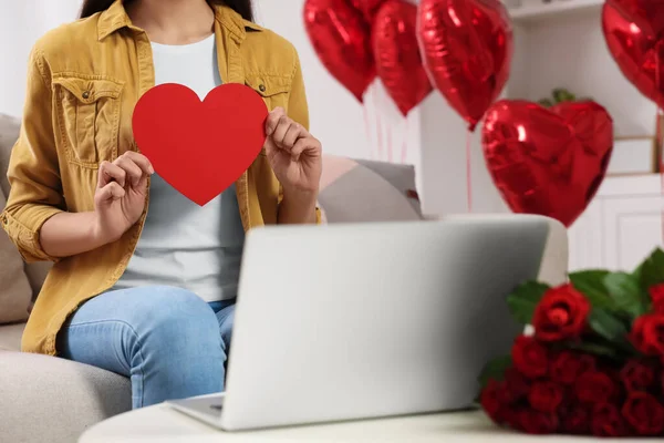 Valentine\'s day celebration in long distance relationship. Woman holding red paper heart while having video chat with her boyfriend via laptop, closeup