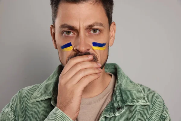 Sad man with drawings of Ukrainian flag on face against light grey background, closeup