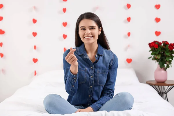 Beautiful young woman showing heart gesture in bedroom, view from camera. Valentine\'s day celebration in long distance relationship