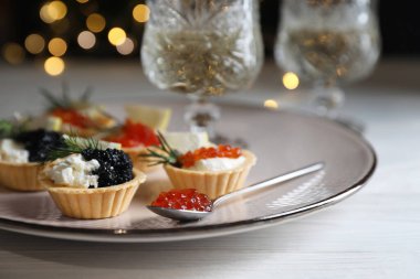Delicious tartlets with red and black caviar served on white wooden table against blurred festive lights, closeup