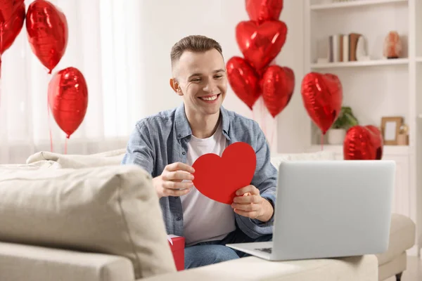 Valentine\'s day celebration in long distance relationship. Man holding red paper heart while having video chat with his girlfriend via laptop at home