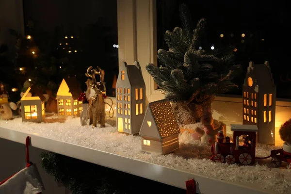 Christmas atmosphere. Beautiful glowing houses, fir trees, artificial snow and toys on window sill indoors