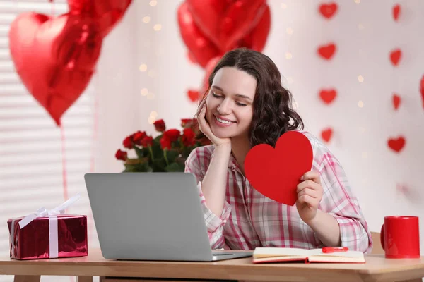 Valentine\'s day celebration in long distance relationship. Woman holding red paper heart while having video chat with her boyfriend via laptop at home