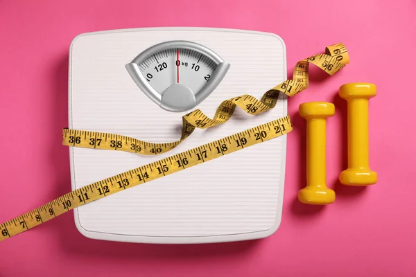 Weight loss concept. Scales, dumbbells and measuring tape on pink background, flat lay