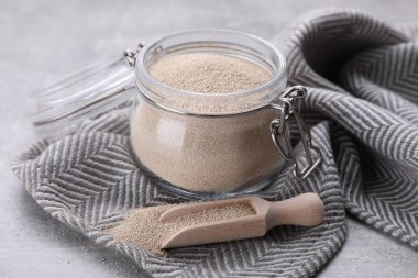 Glass jar and scoop with active dry yeast on light grey table, closeup