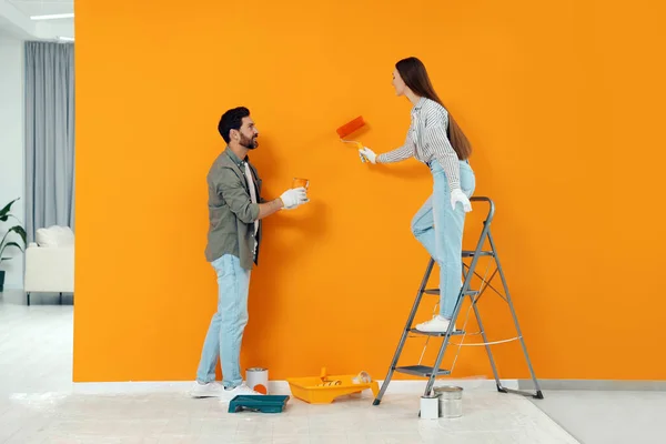 Man holding can of dye and woman painting orange wall indoors. Interior design