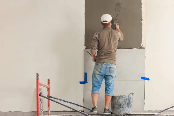 Worker spreading adhesive mix over wall for tile installation indoors, back view