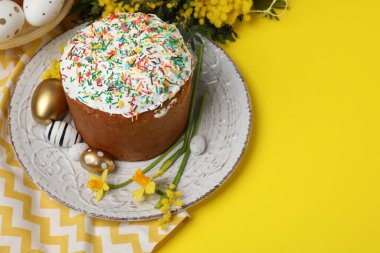 Traditional Easter cake with sprinkles, painted eggs and beautiful spring flowers on yellow background, space for text