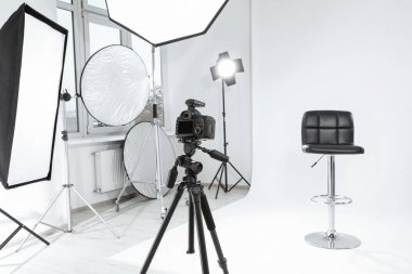 Tripod with camera, bar stool and professional lighting equipment in modern photo studio