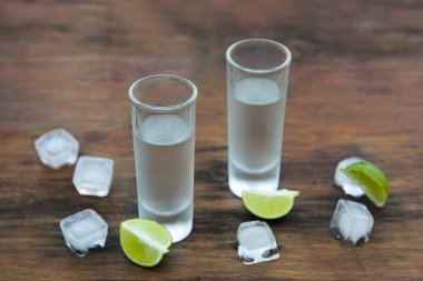 Mexican tequila shots with lime slices and ice cubes on wooden table. Drink made from agave