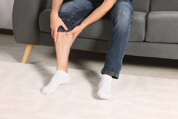 Man suffering from leg pain on sofa indoors, closeup
