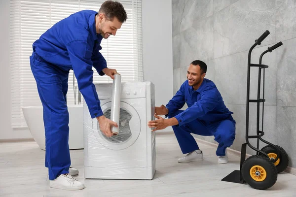 Male movers with stretch film wrapping washing machine in bathroom. New house