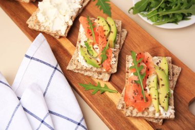 Fresh crunchy crispbreads with cream cheese, salmon, avocado and arugula on beige table