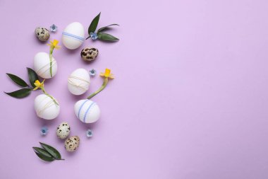 Festively decorated Easter eggs and green leaves on lilac background, flat lay. Space for text