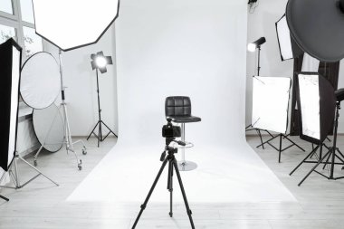 Tripod with camera, bar stool and professional lighting equipment in modern photo studio