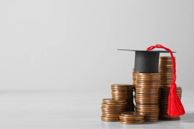 Scholarship concept. Graduation cap and stacked coins on white wooden table, space for text