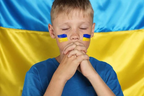 Little boy with clasped hands praying for Ukraine near national flag. No war concept