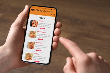 Food delivery service. Man choosing dish from menu on site using smartphone at wooden table, closeup